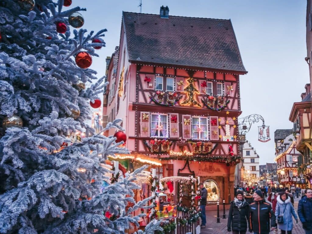 Snow-dusted Christmas tree with red and gold baubles stands in front of a timber-framed building decorated with lights and garlands in a festive European town. People stroll below as part of a charming Christmas market scene.