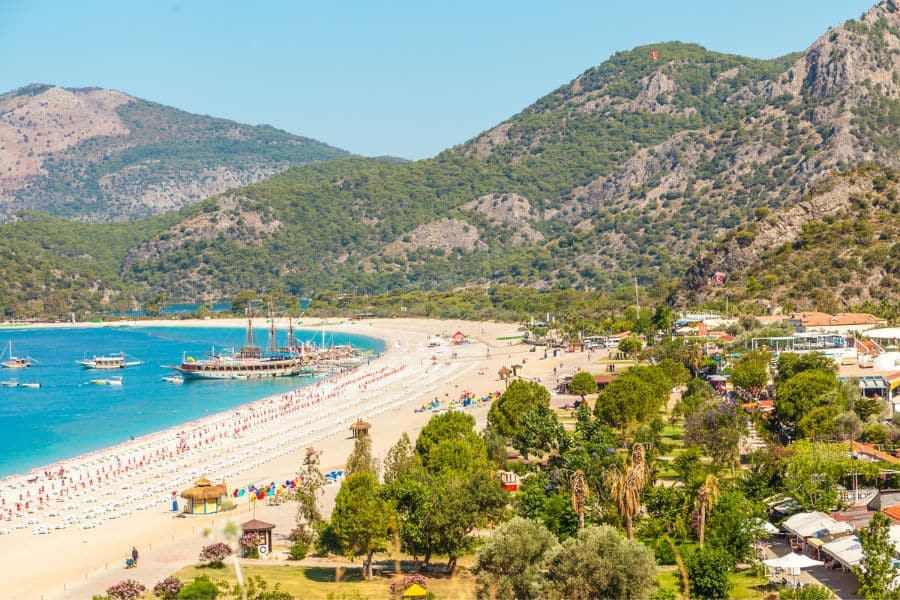 Beautiful sandy beach in Ölüdeniz, Turkey with colorful umbrellas, boats in the bay, and lush green mountains in the background. A peaceful choice for all-inclusive resort getaways combining beach and nature.