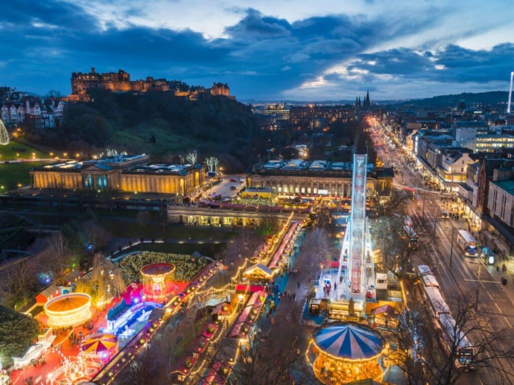 Aerial view of Edinburgh’s Christmas market at dusk, featuring a brightly lit Ferris wheel, carnival rides, and rows of festive stalls beneath Edinburgh Castle. This bustling scene captures the magic of European Christmas markets in the UK.