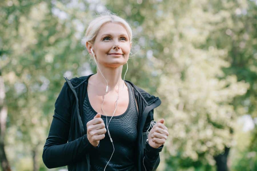 Older woman in athletic wear walking outdoors with earbuds in, looking peaceful and focused. Starting a fitness routine without proper guidance can lead to healthy habit mistakes.