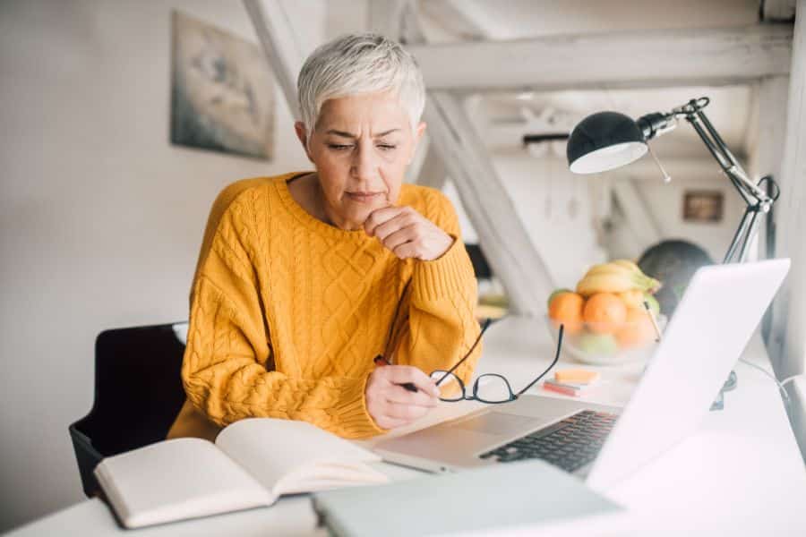 An older woman with short gray hair and a mustard yellow sweater sitting at a bright desk, studying or working on her laptop with books and notes around her. This scene highlights lifelong learning and remote work options for American retirees.