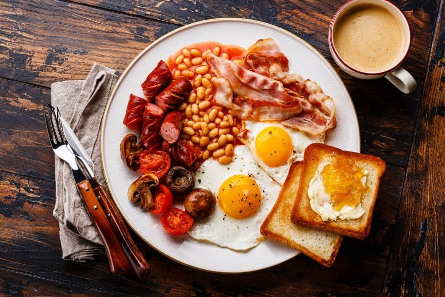 A traditional English breakfast with fried eggs, bacon, sausage, baked beans, toast with butter and marmalade, mushrooms, tomatoes, and coffee. High-fat and highly processed breakfast items are known ingredients that sabotage your memory.