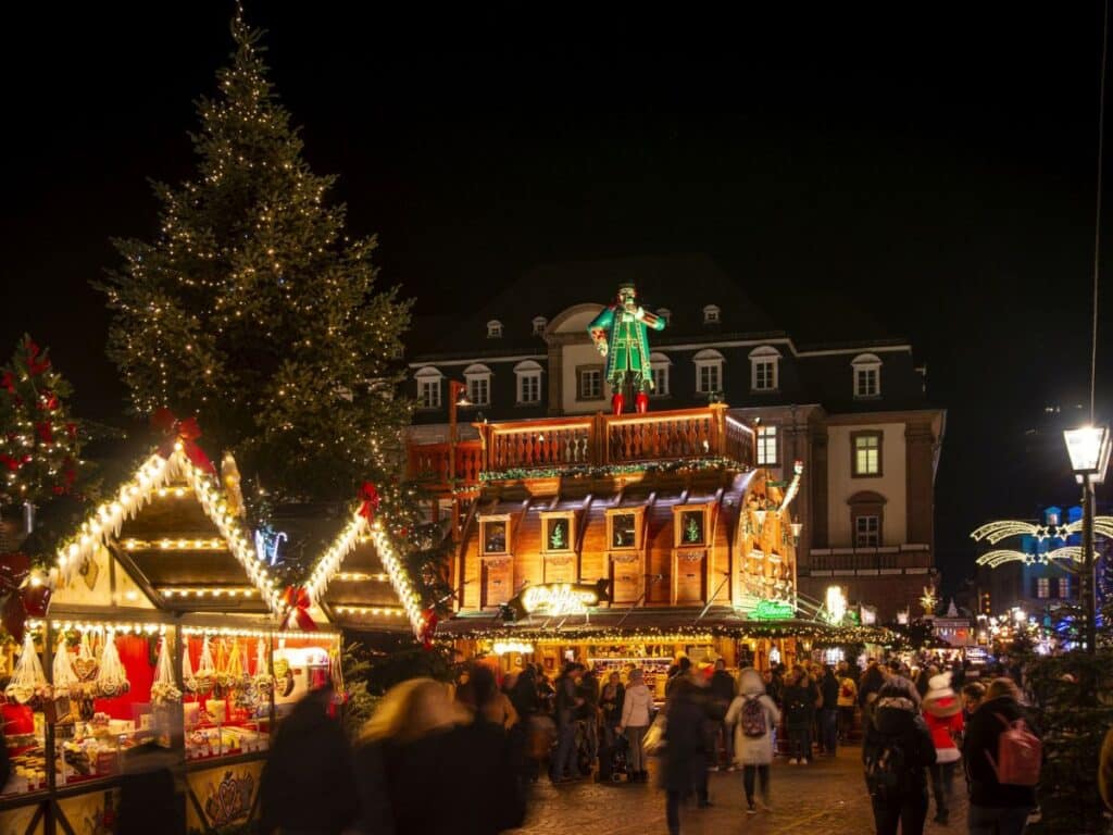 Nighttime view of a Christmas market with lighted stalls and a large Christmas tree, with a traditional wooden building topped by a nutcracker figure. Crowds gather to shop and enjoy the festive spirit found in European Christmas markets.