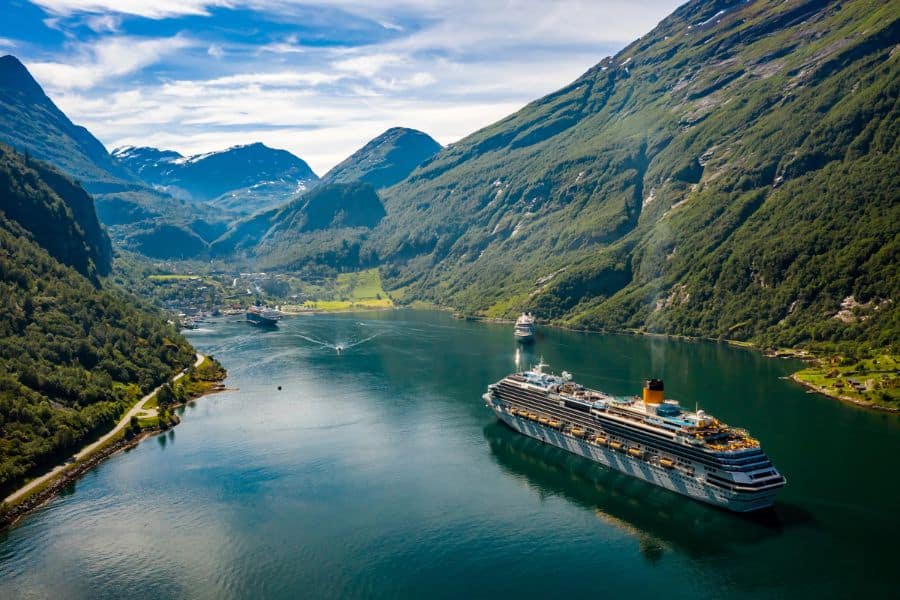A large cruise ship sails through a narrow, scenic fjord surrounded by steep, lush green mountains under a clear sky. This serene setting captures the essence of immersive cruise travel, offering passengers deep connection with nature and remote landscapes.