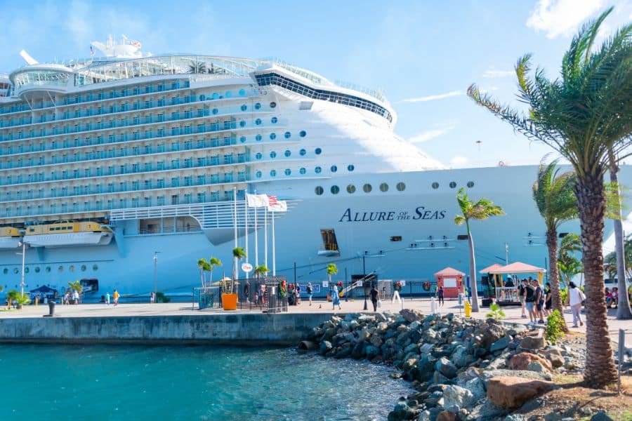 Close-up of the massive Allure of the Seas cruise ship docked at a sunny port, with palm trees and people in the foreground. Highlights the kind of vessel you might board when you pack for a Caribbean cruise.