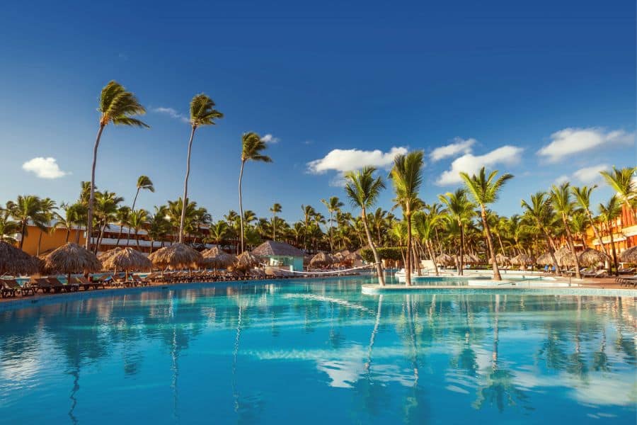 Sparkling blue pool surrounded by palm trees and thatched-roof cabanas under a bright sky at a tropical Caribbean resort. Classic setting for all-inclusive resort getaways offering sun, drinks, and serenity.