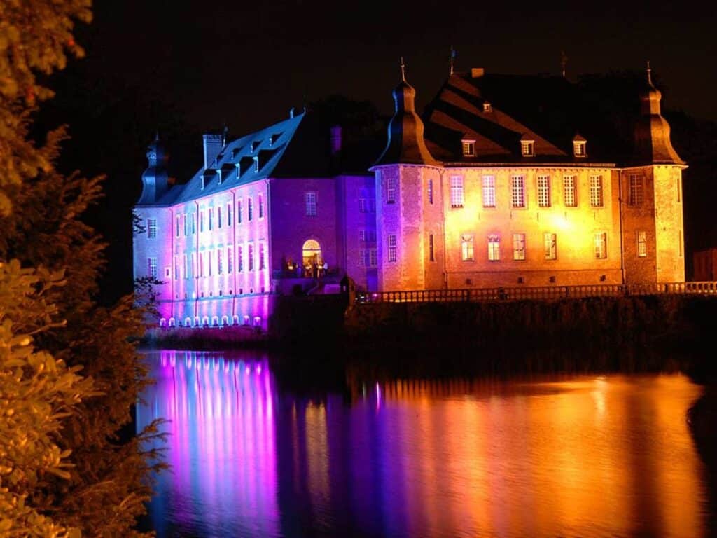 Castle illuminated in bright purple, blue, and golden tones reflecting off a still river at night. Though not a market, the festive lighting echoes the magical ambiance often found near European Christmas market locations.