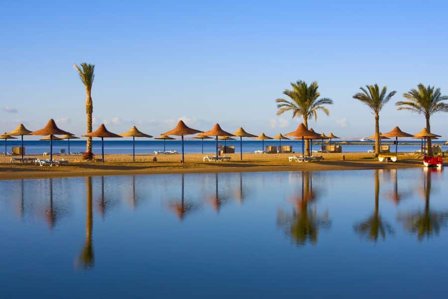 Tranquil beach scene in Hurghada, Egypt with rows of straw parasols, palm trees, and sun loungers reflecting in a calm lagoon. Ideal for relaxing all-inclusive resort getaways on the Red Sea.