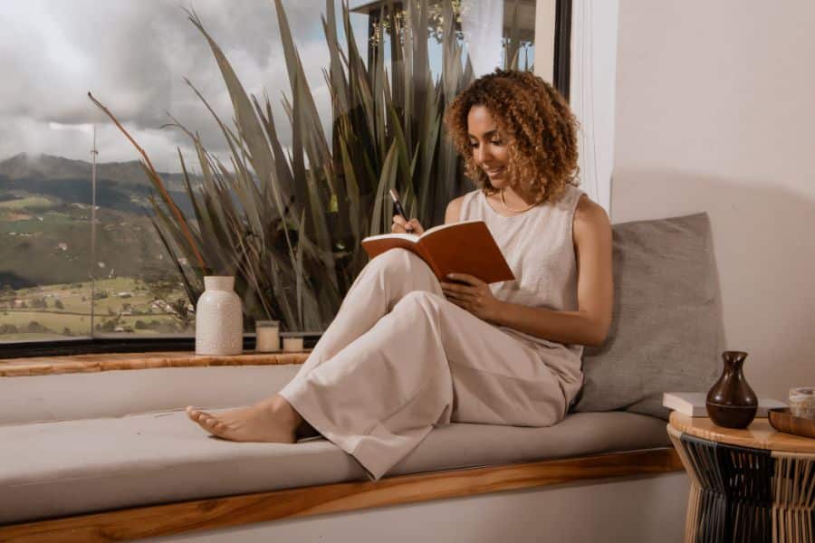 Woman sitting on a window seat journaling in a cozy modern home with lush countryside visible outside. Reflects the relaxed lifestyle and inspiring surroundings of house-sitting in a scenic rural destination.