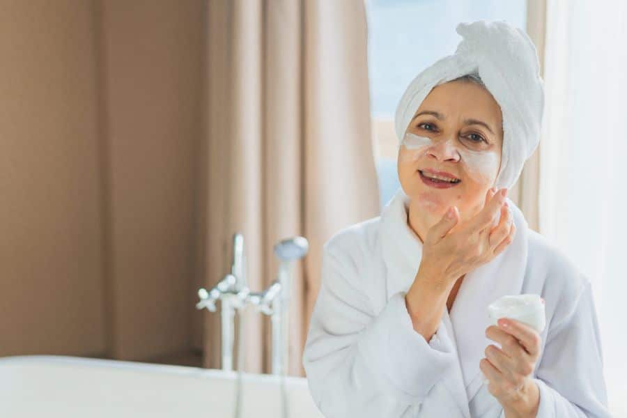 Smiling woman in a bathrobe applying face cream while standing in a bathroom with a towel wrapped around her head. Overusing skincare products is one of the common healthy habit mistakes.