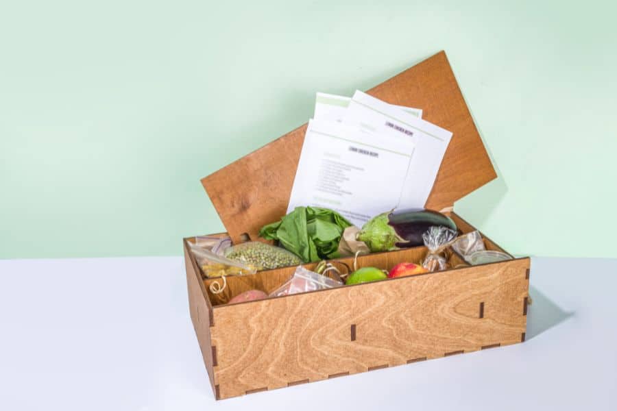 A wooden meal kit box filled with fresh produce like spinach, eggplant, and apples, along with printed recipe cards. Meal delivery services support healthy eating habits for busy or home-based American retirees.