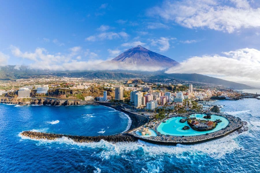 Panoramic view of Puerto de la Cruz, Tenerife with vibrant resort buildings, a lagoon-style pool complex, and Mount Teide rising behind the city. A scenic and volcanic backdrop for European all-inclusive resort getaways.