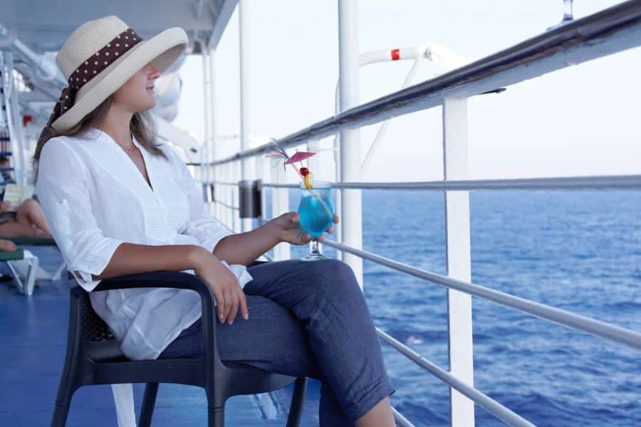 Woman in a sunhat and white blouse relaxing with a blue cocktail on a cruise ship deck overlooking the ocean. Perfect example of why packing light, breezy clothing is key when you pack for a Caribbean cruise.