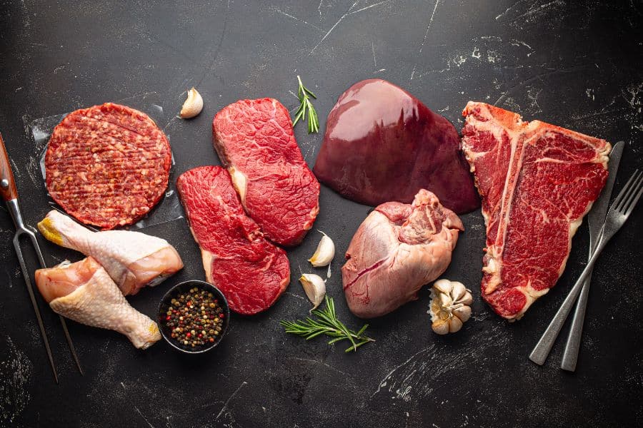 An assortment of raw meats including chicken drumsticks, beef cuts, liver, heart, and a burger patty laid out on a dark surface. Organ meats and fatty cuts may be considered ingredients that sabotage your memory when consumed in excess due to saturated fat.