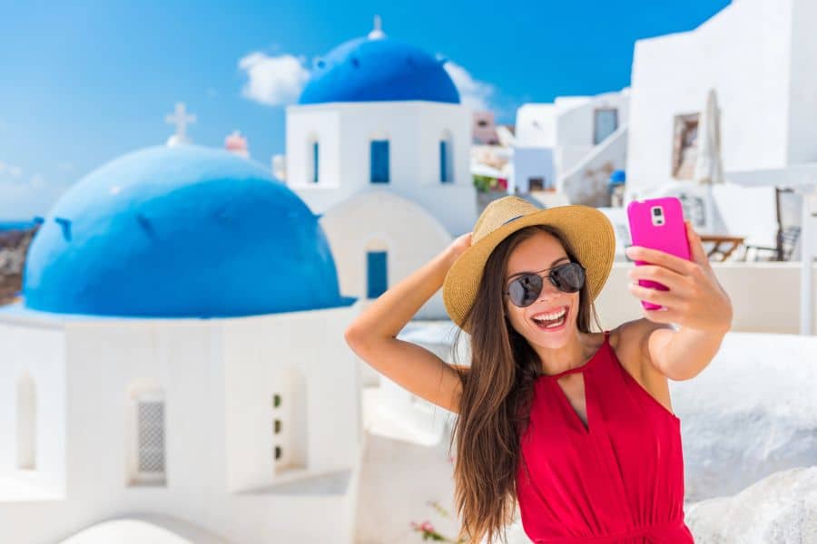 Smiling woman in a red dress takes a selfie in front of iconic blue-domed churches in Santorini, Greece, capturing travel hacks for Instagram-worthy moments.