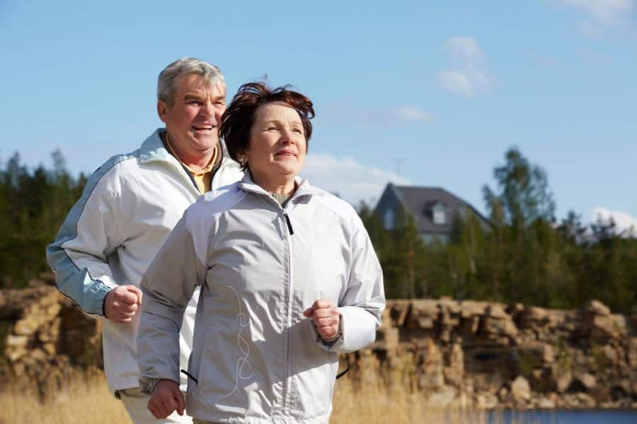 Mature couple jogging near a lake with forest and house in the background, emphasizing outdoor fitness as a lasting exercise trend.