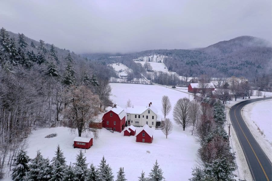 A white farmhouse and red barns sit nestled in a snowy Vermont valley, surrounded by frosted trees and rolling hills. A quintessential rural scene often encountered during cozy winter RV trips through New England.