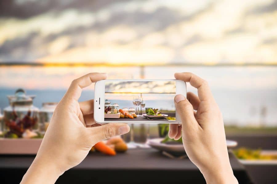 Hands taking a photo of a seaside dinner table using a smartphone, with wine glasses and plated dishes in focus against a sunset backdrop. Captures the essence of dining in the age of Instagram, blending food with scenic storytelling.