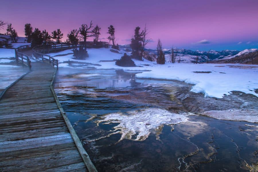 A wooden boardwalk winds across a steamy, snow-covered geothermal area at sunrise, with pastel pink and purple skies above. This Yellowstone scene showcases the surreal landscapes visited during winter RV trips.