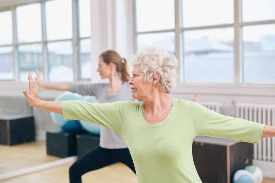 An older woman with gray hair wearing a light green top, holding a yoga pose in a bright fitness studio alongside a younger woman. Group wellness classes are a popular way for American retirees to stay active and social.