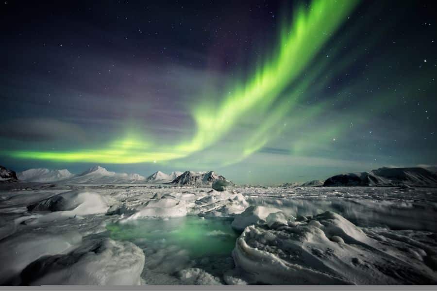 Bright green aurora borealis dances over a snow-covered Arctic landscape with jagged mountains in the distance. One of the best places to see the Northern Lights in a remote, icy wilderness.