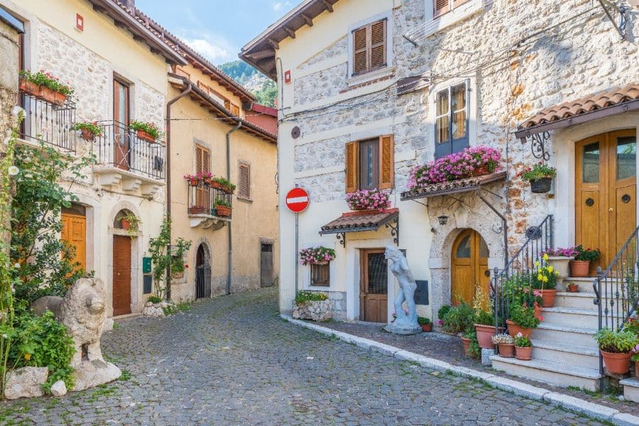 A quiet cobblestone street in Abruzzo, Italy, lined with rustic stone buildings and colorful flower boxes under sunny skies. This peaceful village scene reflects the charm of slow travel destinations in Europe.