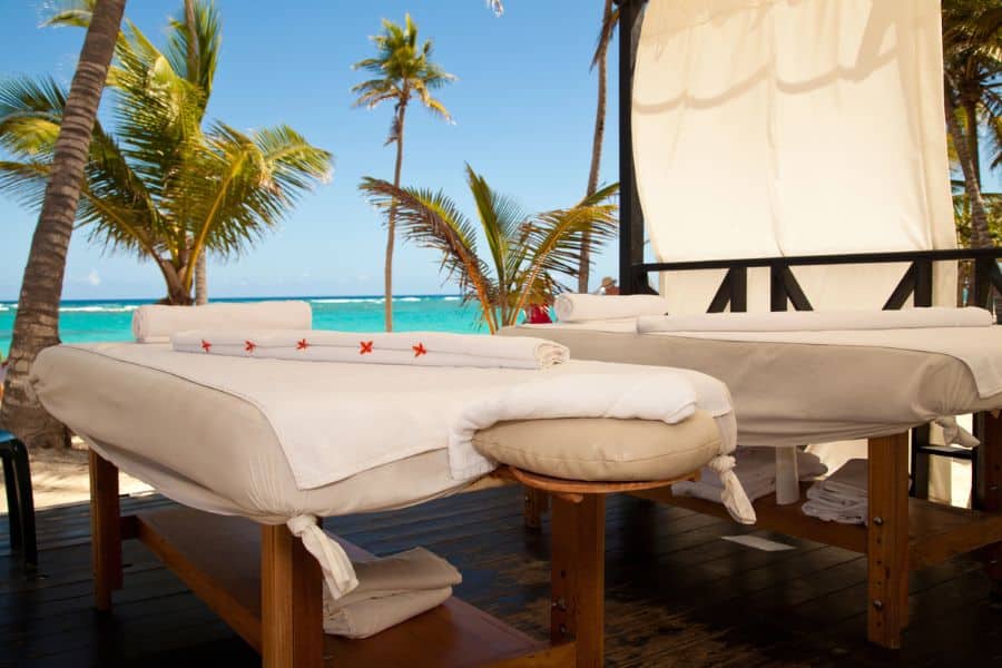 Two massage tables with rolled towels and starfish accents sit under a canopy, facing a turquoise ocean surrounded by swaying palm trees. This tranquil setup captures the essence of Caribbean luxury travel, where beachside spa experiences offer ultimate relaxation in paradise.