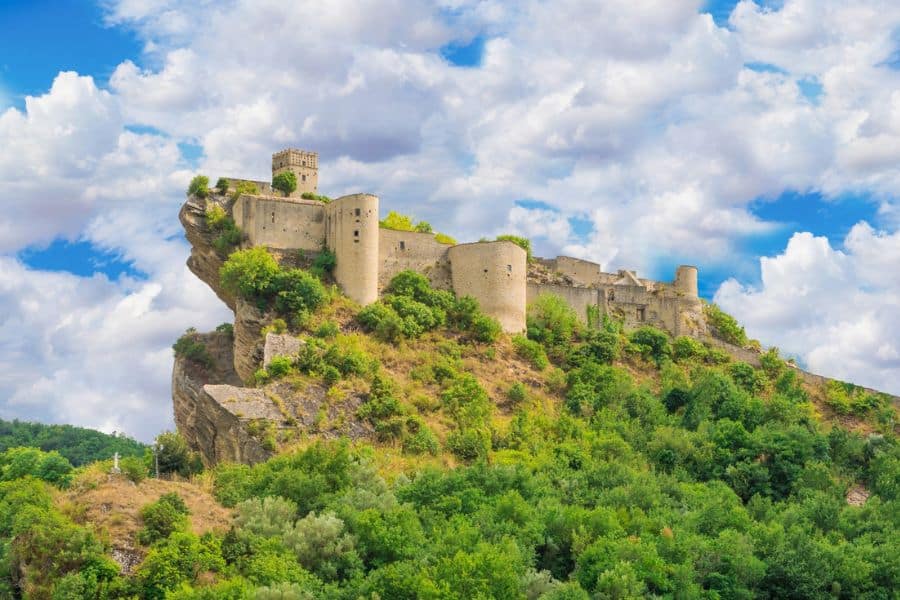 A dramatic hilltop fortress perched above lush green hills and cliffs in Abruzzo, Italy, with thick stone walls and a watchtower under a cloudy sky. This historic site embodies the timeless allure of slow travel destinations in Europe.