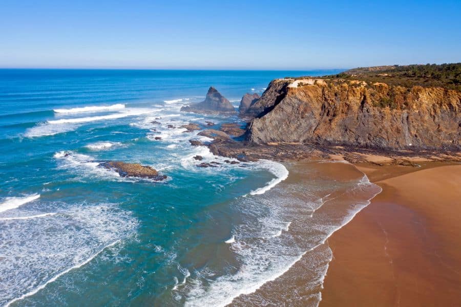 A sweeping view of Alentejo’s rugged coastline with golden cliffs, gentle surf, and an empty sandy beach meeting turquoise waters. This tranquil seascape highlights why Alentejo is one of the best slow travel destinations in Europe.