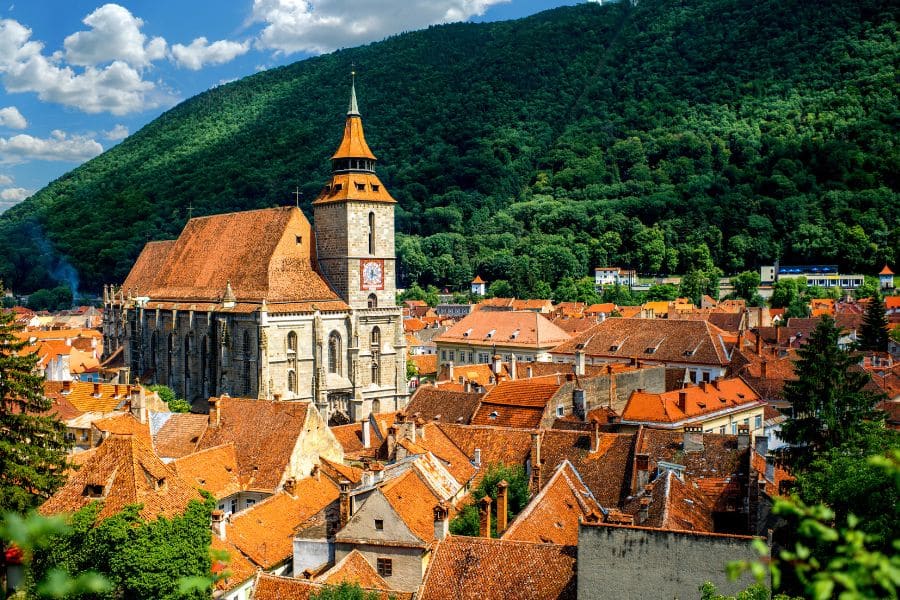 A sweeping view of Brasov, Romania, featuring the Black Church with its clock tower surrounded by orange-roofed houses and forested hills. A perfect example of towns in Europe where you can live for less while enjoying medieval charm.