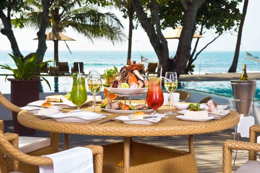 A beachfront dining table is elegantly set with tropical cocktails, white wine, and a lavish seafood platter featuring lobster, prawns, and shellfish, framed by palm trees and ocean views. This image reflects the indulgent experience of Caribbean luxury travel, combining fine dining with a serene coastal ambiance.