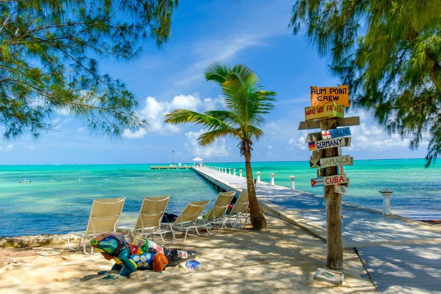 A white wooden pier stretches out over calm turquoise waters, with lounge chairs, palm trees, and a whimsical signpost pointing to destinations like Jamaica and Cuba. This peaceful beachscape captures the charm and vibrant character of Caribbean luxury travel.