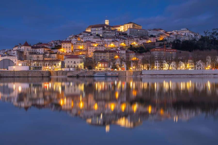 A scenic evening view of Coimbra, Portugal, with warm city lights reflecting on the calm river below. Portugal is one of the countries that welcome Americans with open arms.