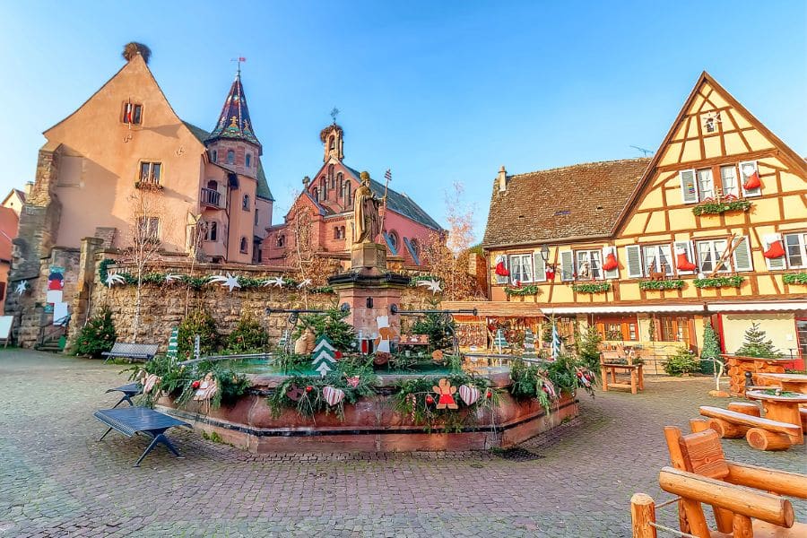 A festive square decorated for the holidays in one of the quaint villages in Europe, featuring a statue, timber-framed buildings, and a peaceful church.