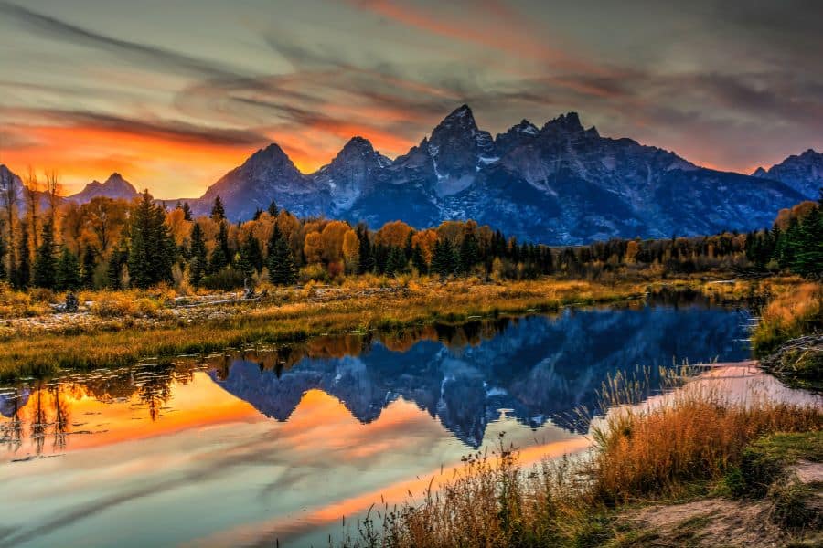 Sunset over Grand Teton National Park, with mountain silhouettes mirrored in a reflective river surrounded by golden autumn trees. The area’s popularity leads to traffic jams and packed viewpoints, often placing it among national parks to avoid during busy months.