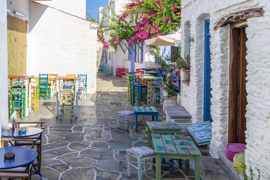 A colorful alleyway in Kythnos, Greece, with stone-paved paths, rainbow-painted tables and chairs, and blooming bougainvillea climbing whitewashed walls. This charming setting exemplifies the joy of exploring slow travel destinations in Europe.