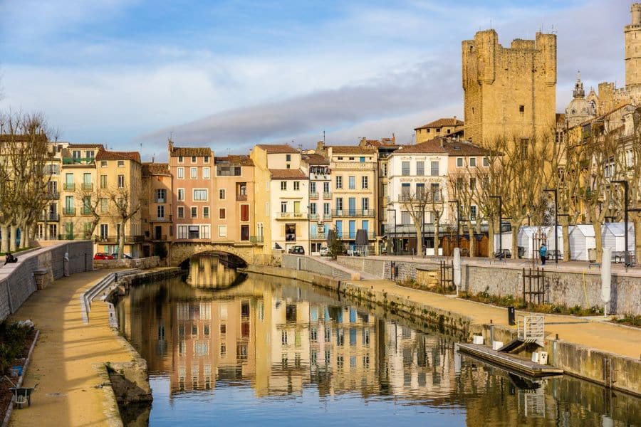Pastel-colored buildings reflect in a peaceful canal in Languedoc, France, with a stone bridge and a medieval tower rising in the background. The serene riverside scene makes it a classic example of slow travel destinations in Europe.
