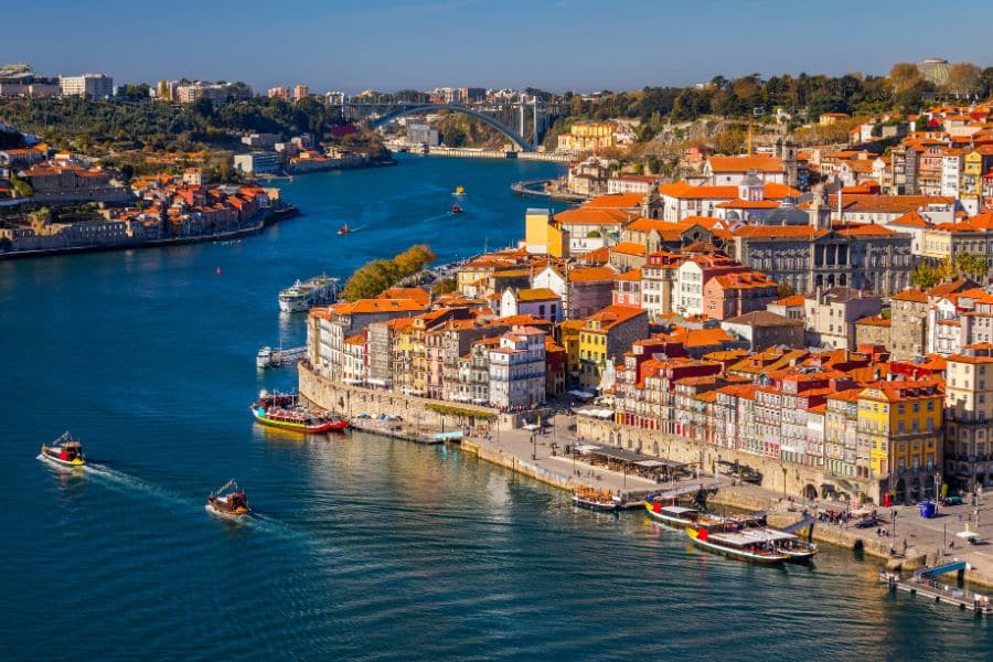 Colorful riverside buildings and traditional boats line the Douro River in Porto, Portugal, under a sunny sky with arched bridges in the distance. A lively, scenic choice among towns in Europe where you can live for less.
