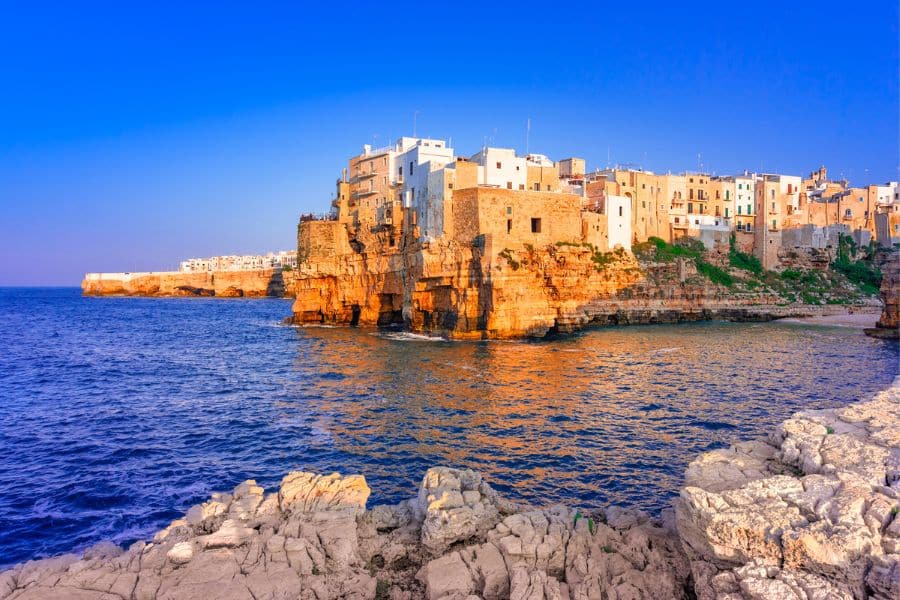 A dramatic cliffside town built over rugged rocks glows orange in the evening sun as it meets the deep blue Adriatic Sea. This dramatic seaside setting is one of many found in Beautiful Italian Regions.