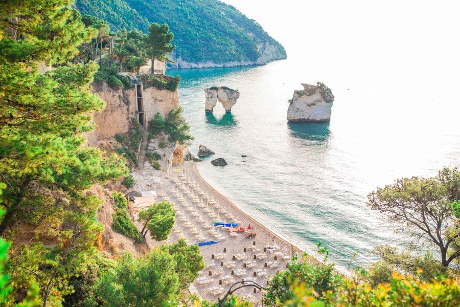 A quiet beach framed by cliffs and pine trees features rows of sun loungers facing sea stacks in the water. This peaceful shoreline is part of the coastal magic in Beautiful Italian Regions.