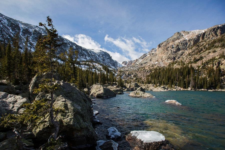 Rocky shoreline of an alpine lake in Rocky Mountain National Park, surrounded by snow-dusted peaks and evergreen trees under a bright blue sky. Despite the scenery, overcrowded trails and parking issues make this one of the national parks to avoid during peak travel times.