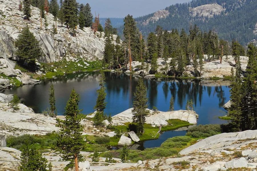 Peaceful lake nestled among granite cliffs and pine trees in Kings Canyon National Park, reflecting a clear sky and untouched wilderness. While beautiful, its remote trails and minimal visitor services place it on lists of national parks to avoid for casual tourists.