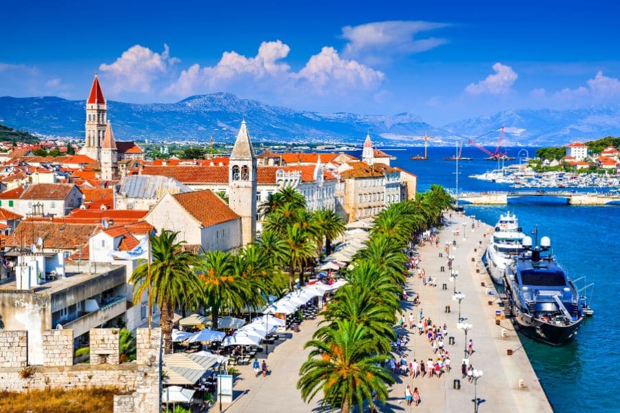 Palm-lined promenade along the Adriatic Sea with medieval bell towers and white stone buildings in Split, Croatia. This coastal paradise is one of the towns in Europe where you can live for less without sacrificing beauty.