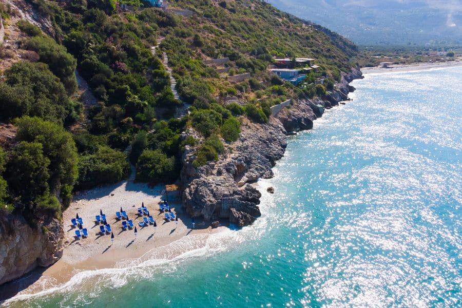 An aerial view of a secluded beach on the Albanian Riviera with rows of blue umbrellas, turquoise water, and rocky cliffs covered in greenery. This untouched coastline showcases the hidden beauty of slow travel destinations in Europe.