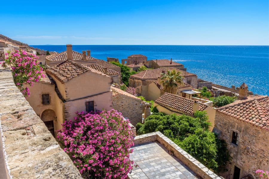 Stone-roofed houses and vibrant bougainvillea bloom along a sunlit coastal village in the Peloponnese, Greece, overlooking the deep blue sea. The laid-back Mediterranean vibe captures the essence of slow travel destinations in Europe.