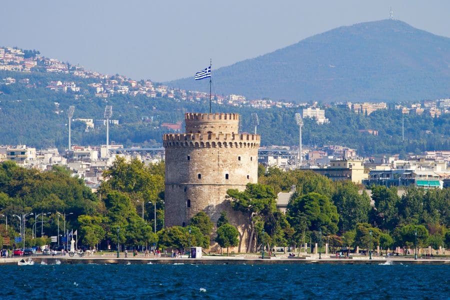 The White Tower of Thessaloniki stands tall by the Aegean Sea, with a Greek flag waving above and a cityscape backed by hills. A historical yet budget-friendly highlight among towns in Europe where you can live for less.