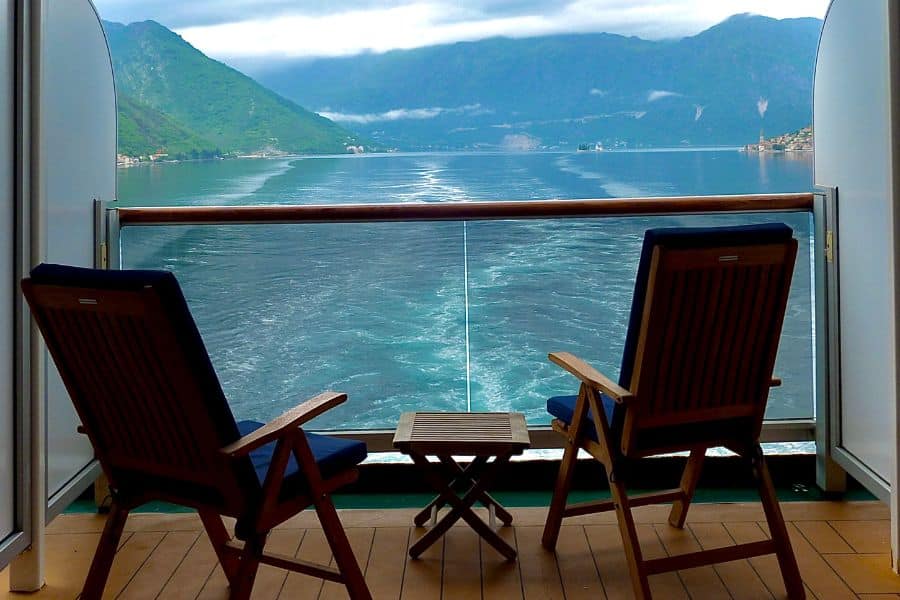 Two wooden deck chairs on a private cruise ship balcony overlook a calm sea and green mountains, capturing the peaceful lifestyle many imagine when they choose to Live on a Cruise Ship.