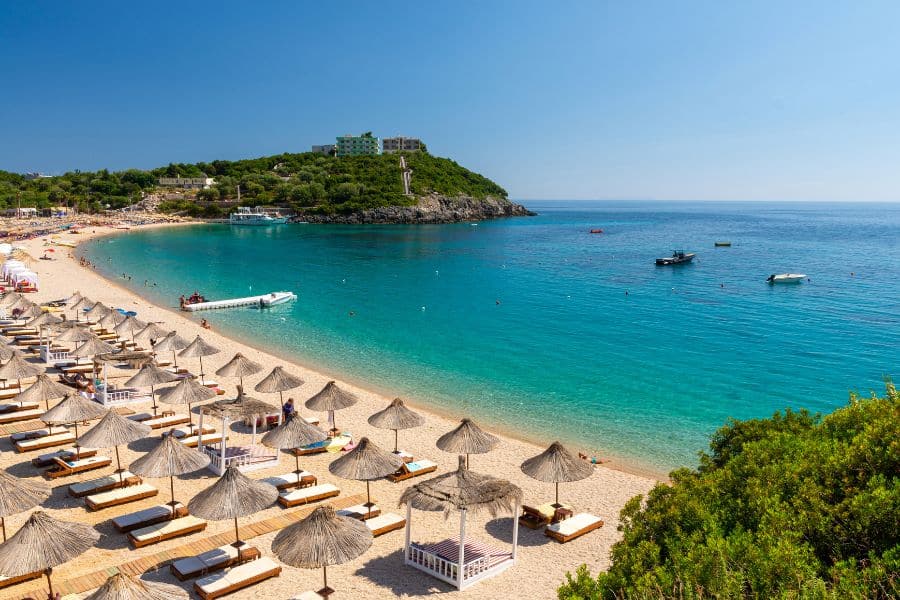 Golden sand beach lined with straw umbrellas and lounge chairs faces calm, clear blue water under a bright sky. A perfect Mediterranean setting to escape the winter blues in January and soak up the sun.