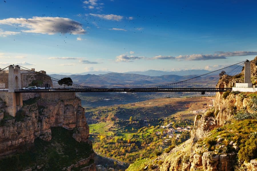 A dramatic suspension bridge stretches across a deep gorge, connecting two cliffs with a sweeping valley below and mountains in the distance. A unique off-season destination for those looking to escape the winter blues in January with scenic views.