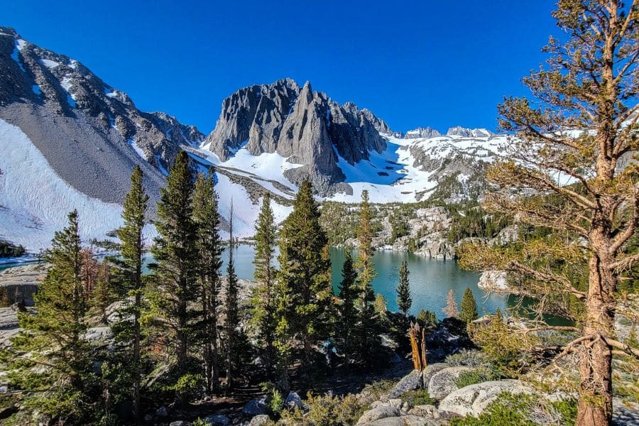Stunning alpine view of jagged peaks, snow patches, and a turquoise lake surrounded by pine trees in the Eastern Sierra, one of America’s Best-Kept Hiking Secrets for mountain lovers.